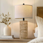 Woven rattan table lamp lit on a dresser beside a cream vase of dried grasses