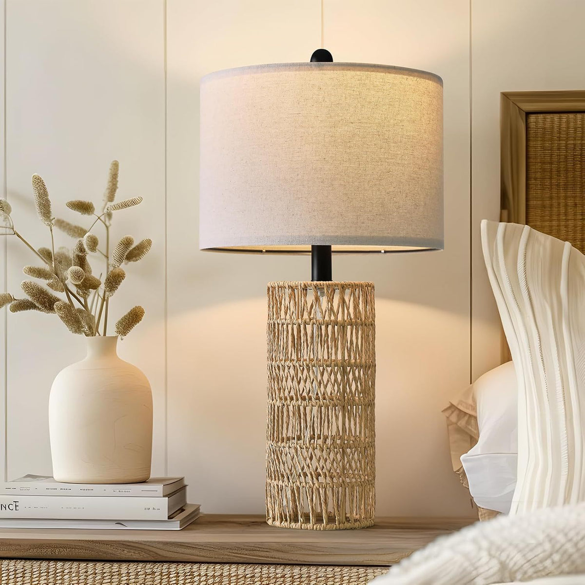 Woven rattan table lamp lit on a dresser beside a cream vase of dried grasses
